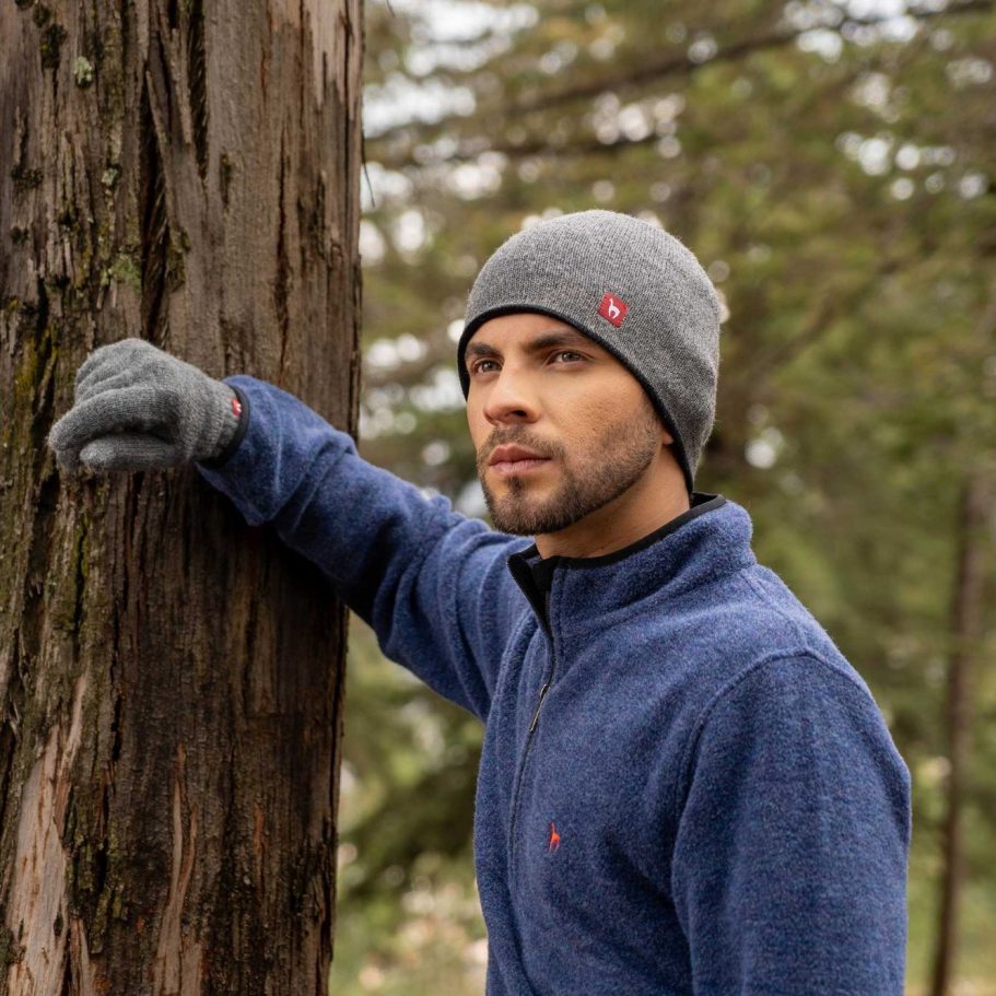 Mann in Alpaka Pullover und Alpaka Mütze lehnt an einem Baum im Wald.