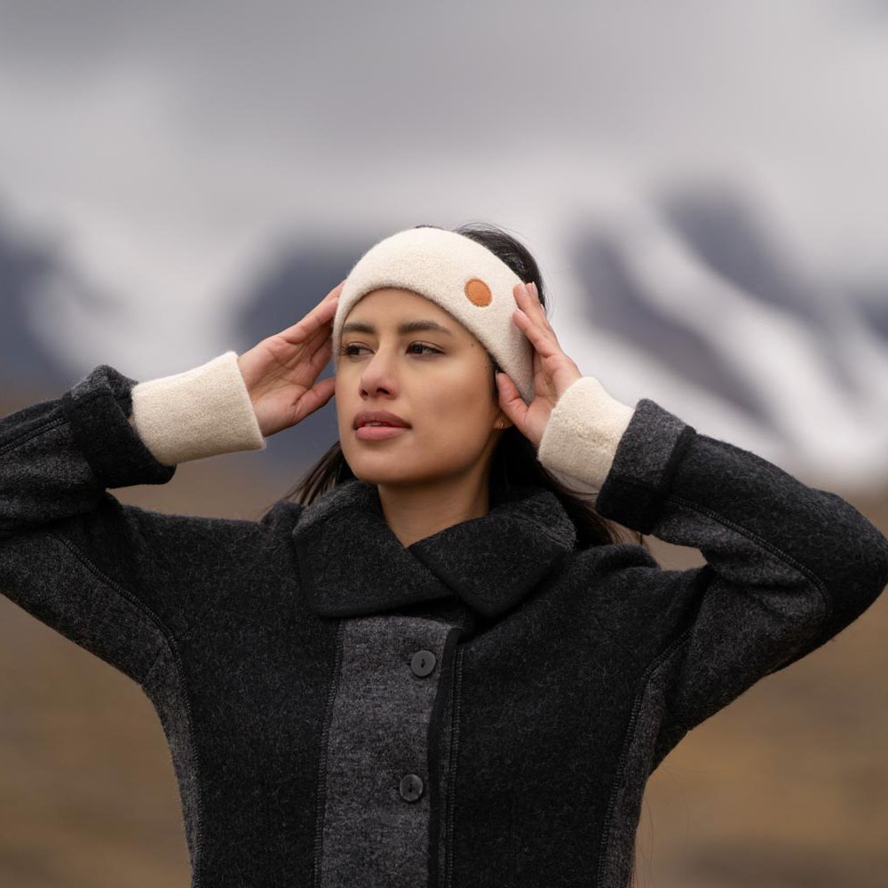 Frau trägt ein Alpaka Stirnband und einen Mantel, mit bergiger Landschaft im Hintergrund.