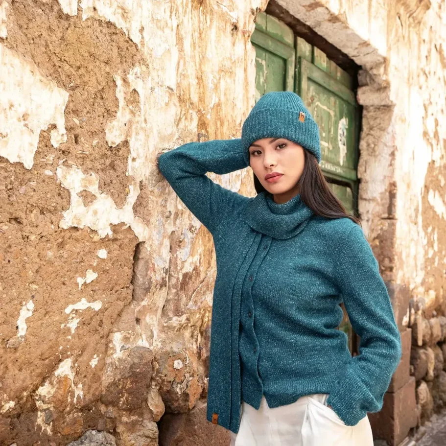 Frau in blauer Alpaka Strickjacke und Alpaka Mütze vor einer rustikalen Wand mit grüner Tür.