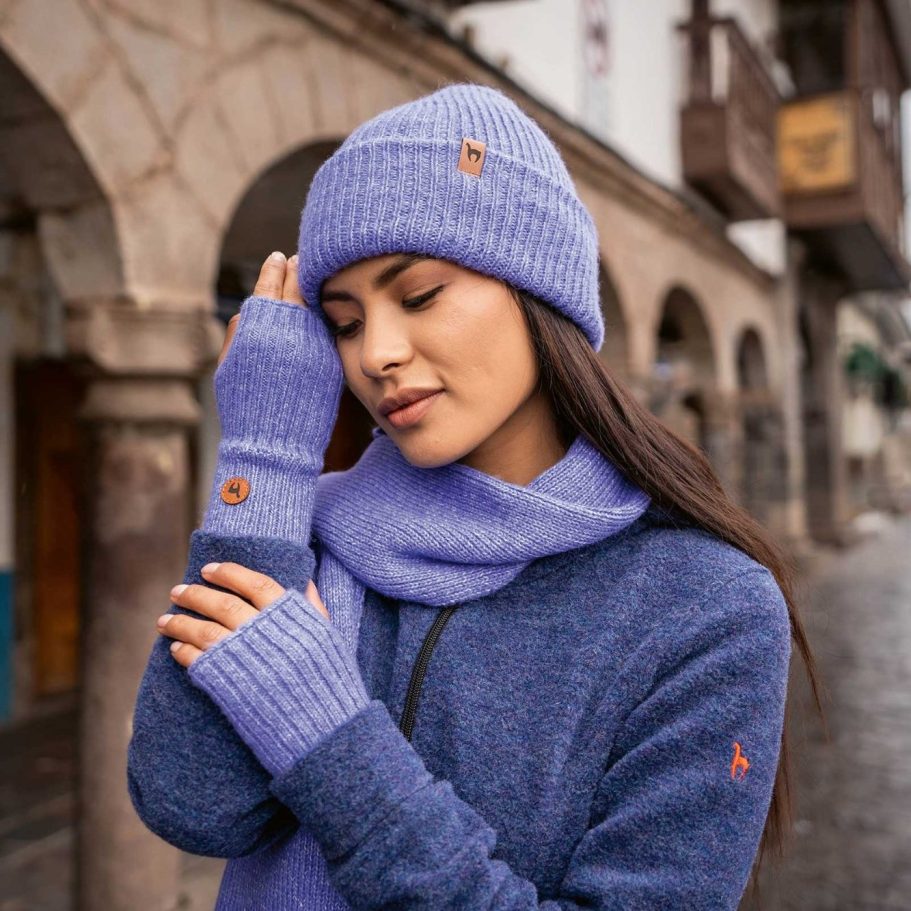 Junge Frau trägt blauen Alpaka Pullover, Alpaka Mütze und Alpaka Schal, steht in einer Stadtstraße.